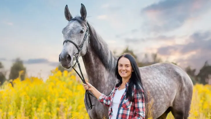 Beautiful woman smiling and holding a dapple gray horse by the reins in a picturesque flowery field