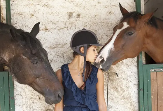 Young woman with a horse riding hat on standing between two horses in their stables