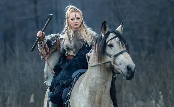 Woman dressed as a barbarian holding an axe while riding a palomino horse in a movie