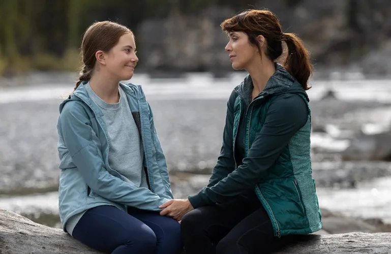 Katie Fleming Morris and Lou Fleming sitting on a log by a river holding hands and smiling at each other in Heartland season 16