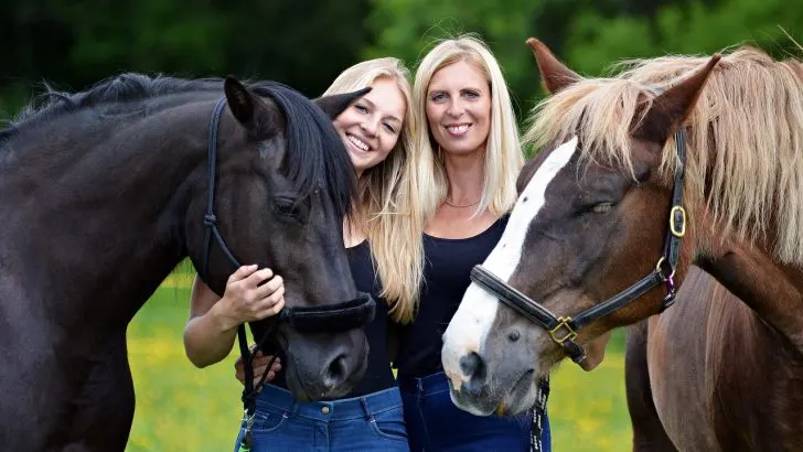 Two blonde hair women standing side by side both with a horse on either side of them